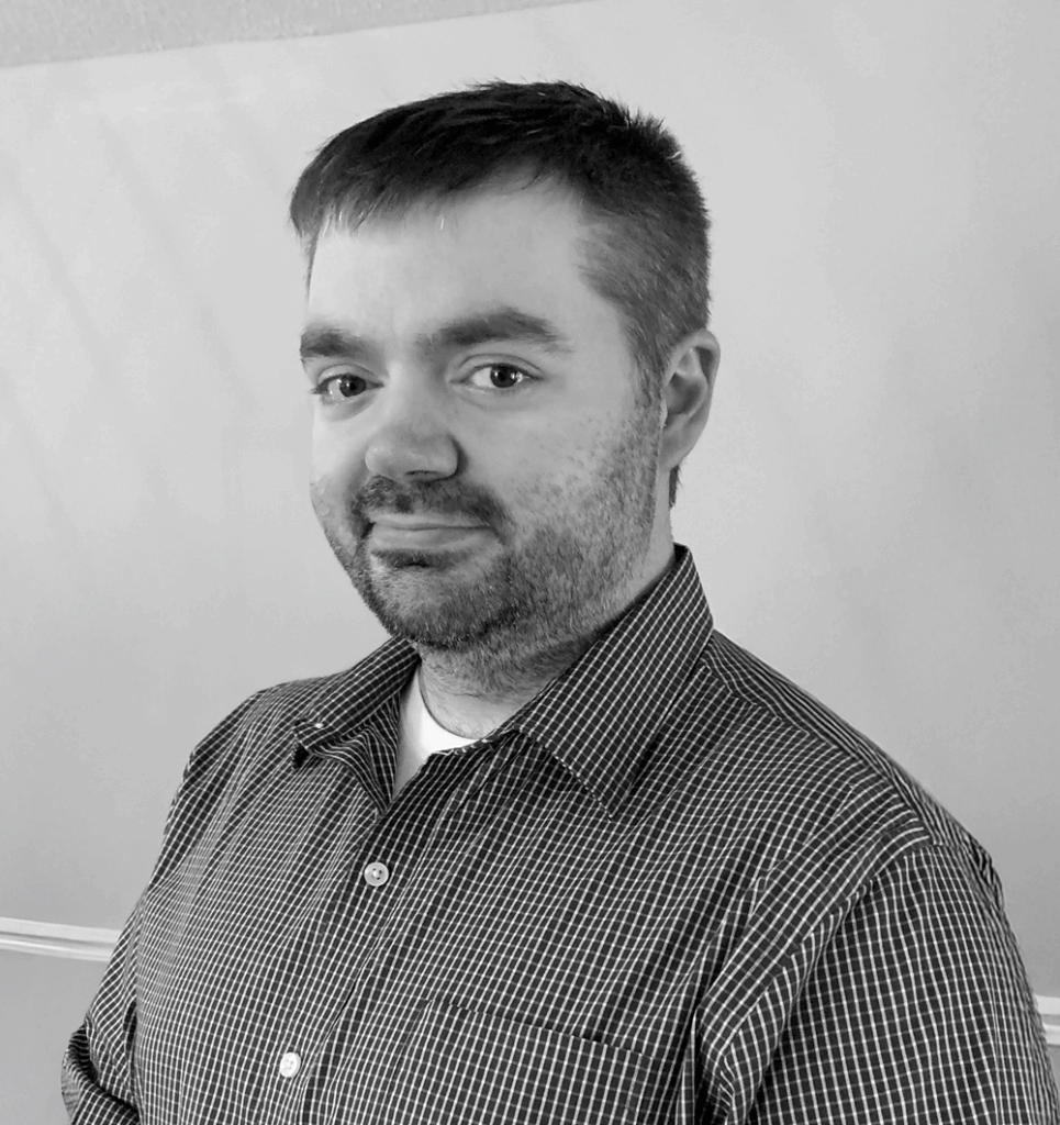 Portrait of James Cody Phenis, a man with short hair and a beard, wearing a checkered button-up shirt, standing indoors with hands clasped in front of him