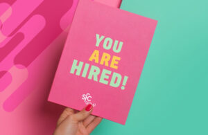 A hand with red nail polish holds a bright pink card that reads "YOU ARE HIRED!" in green and yellow block letters. The card also features a small "sfc group" logo in white. The background is split diagonally, with a darker pink on the left and a teal on the right, and includes some rounded pink shapes.